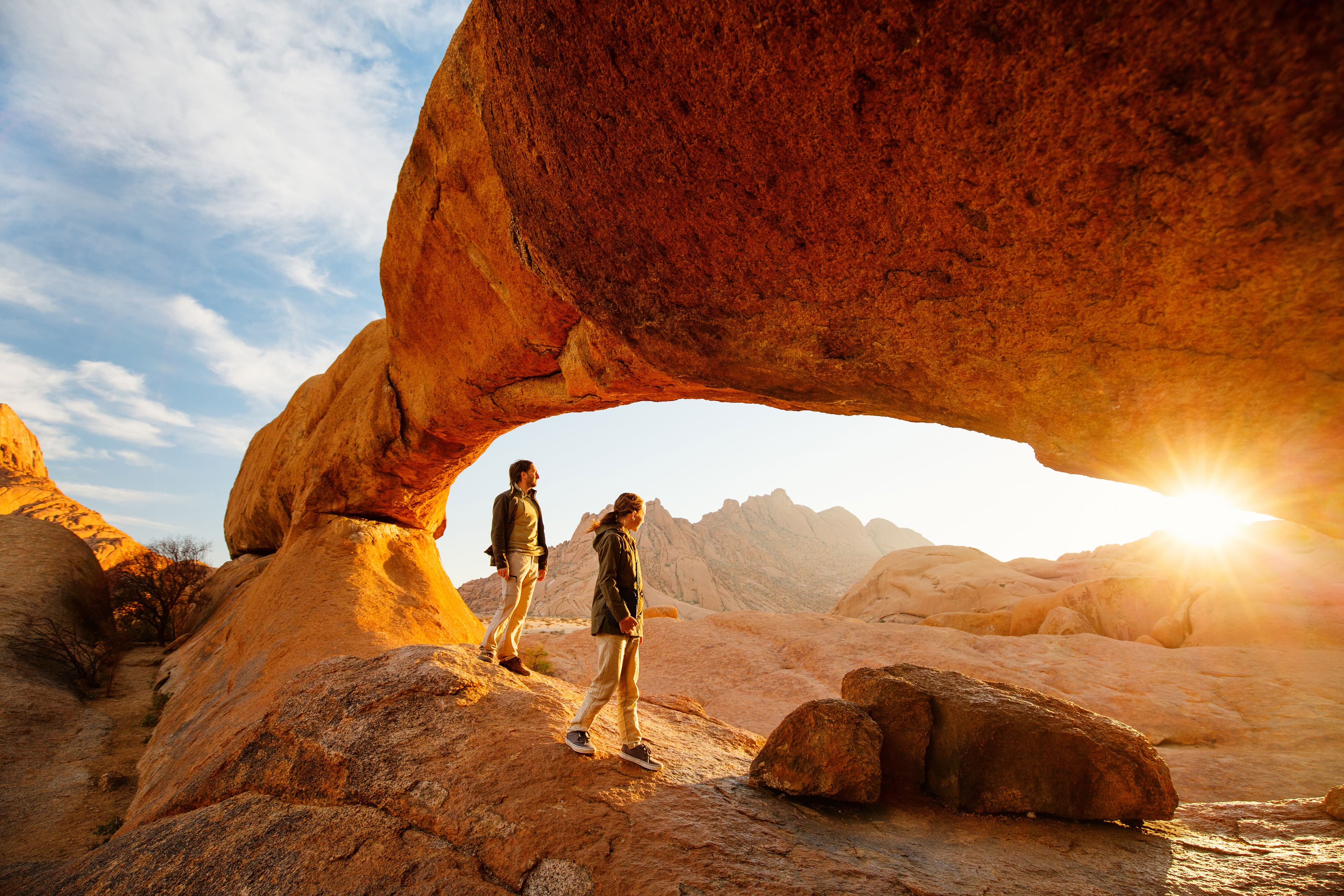 Man and woman hiking below a stone arch at sunrise